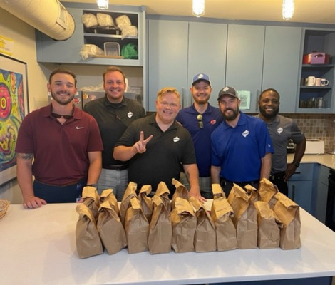 Group of six adult men post in kitchen with blue cabinets behind them. Twenty sack lunches in brown bags are on white counter in front of them.