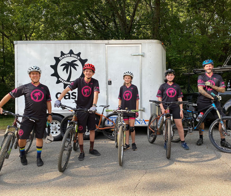 Two adults bookend three youth, all are on bikes, wearing helmets, and dressed in black biking attire with a hot pink logo for the Oasis Bike Workshop. Behind them is a white trailer with the logo and trees.