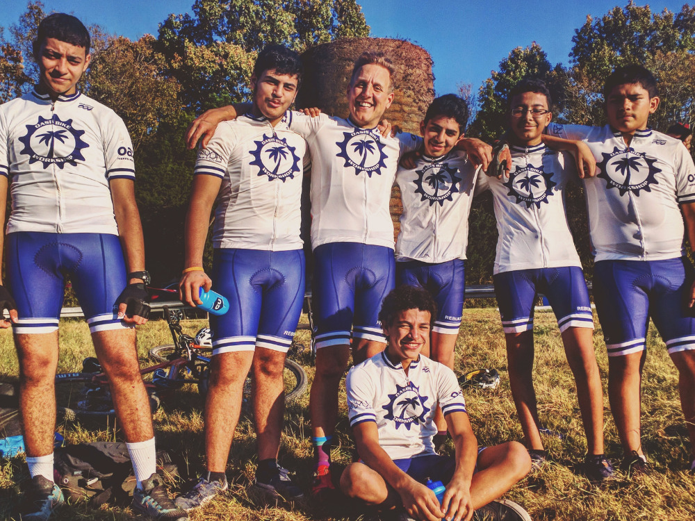 Five youth and one adult are standing, wearing biking attire of white shirts and blue shorts. One youth is sitting on the ground in front of them.